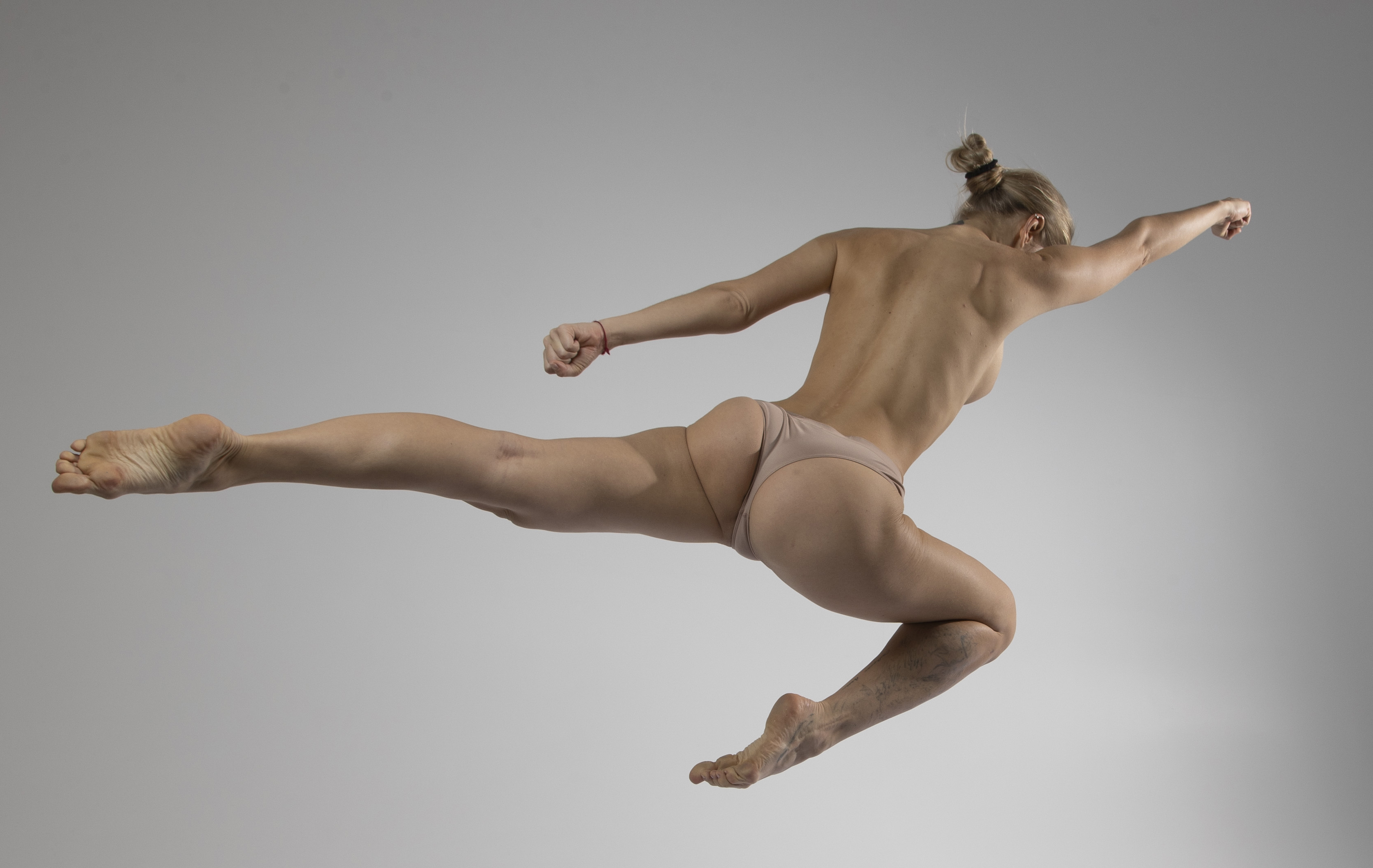 a nude woman wearing underwear is captured midair in a dynamic jumping pose. she is blonde with her hair in a bun. the background is a plain studio backdrop. her physique suggests fitness and athleticism.