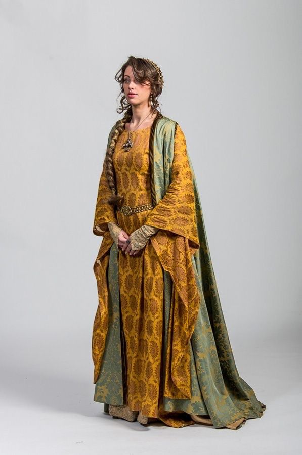 a woman wearing a long yellow gown with a paisley pattern and a green cape stands in front of a white background. she has a long braid, and her hair is also adorned with jewelry. she is also wearing a necklace and a belt. her hands are clasped together.