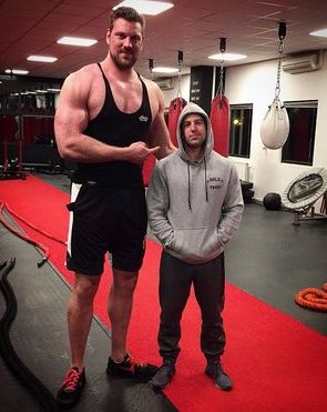 the image shows a photograph of two men in a gym. one man is extraordinarily tall and muscular, likely a bodybuilder or strongman, wearing a black tank top and black shorts.  he stands significantly taller than the other man, who is of average height and wearing a grey hoodie and sweatpants. the taller man is pointing at the shorter man.  the gym setting includes heavy bags and other workout equipment. a red mat or carpet is visible on the gym floor. the overall contrast in height between the two men is the striking feature of the image.