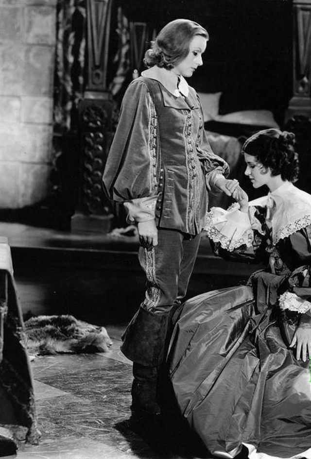 a woman dressed in a historical costume looks down at a man in a similar costume while holding his hand. they are both on stage, and the photo is likely a still from a film.