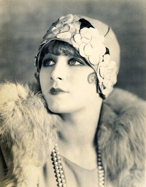 a black and white portrait of a woman in a vintage 1920s flapper style. she is wearing a hat with a floral design, a fur coat, and a pearl necklace. she has dark makeup on her eyes and lips. the image has a retro feel and captures the fashion of the era.