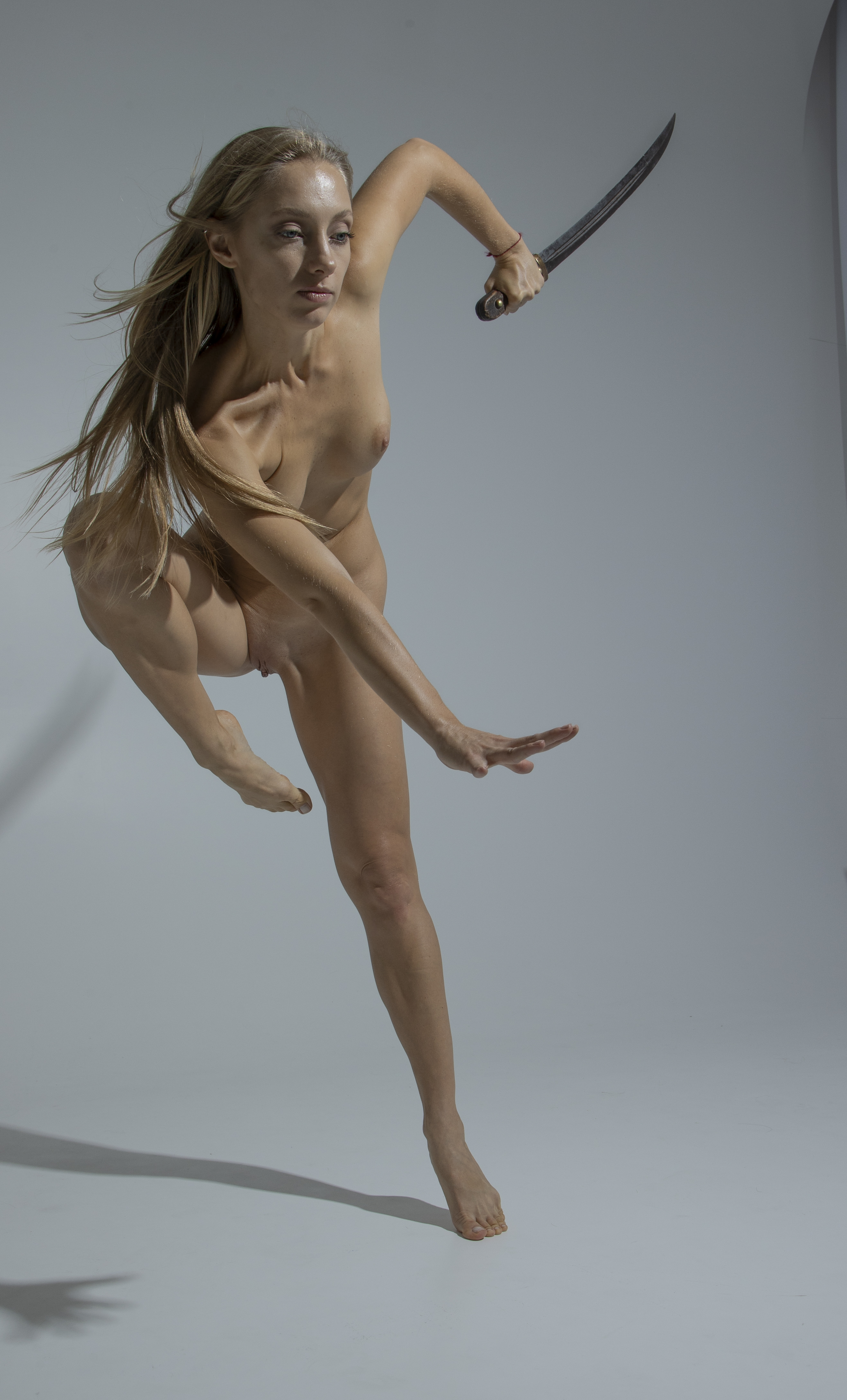 a fullbody studio shot of a nude woman with blonde hair, captured midjump, wielding a sword. she is barefoot, with a slender build, and her pose suggests a dynamic, warriorlike stance. the background is a plain, light gray tone, emphasizing the figure's form and movement. her hair flows to the side and her face is looking to the side.