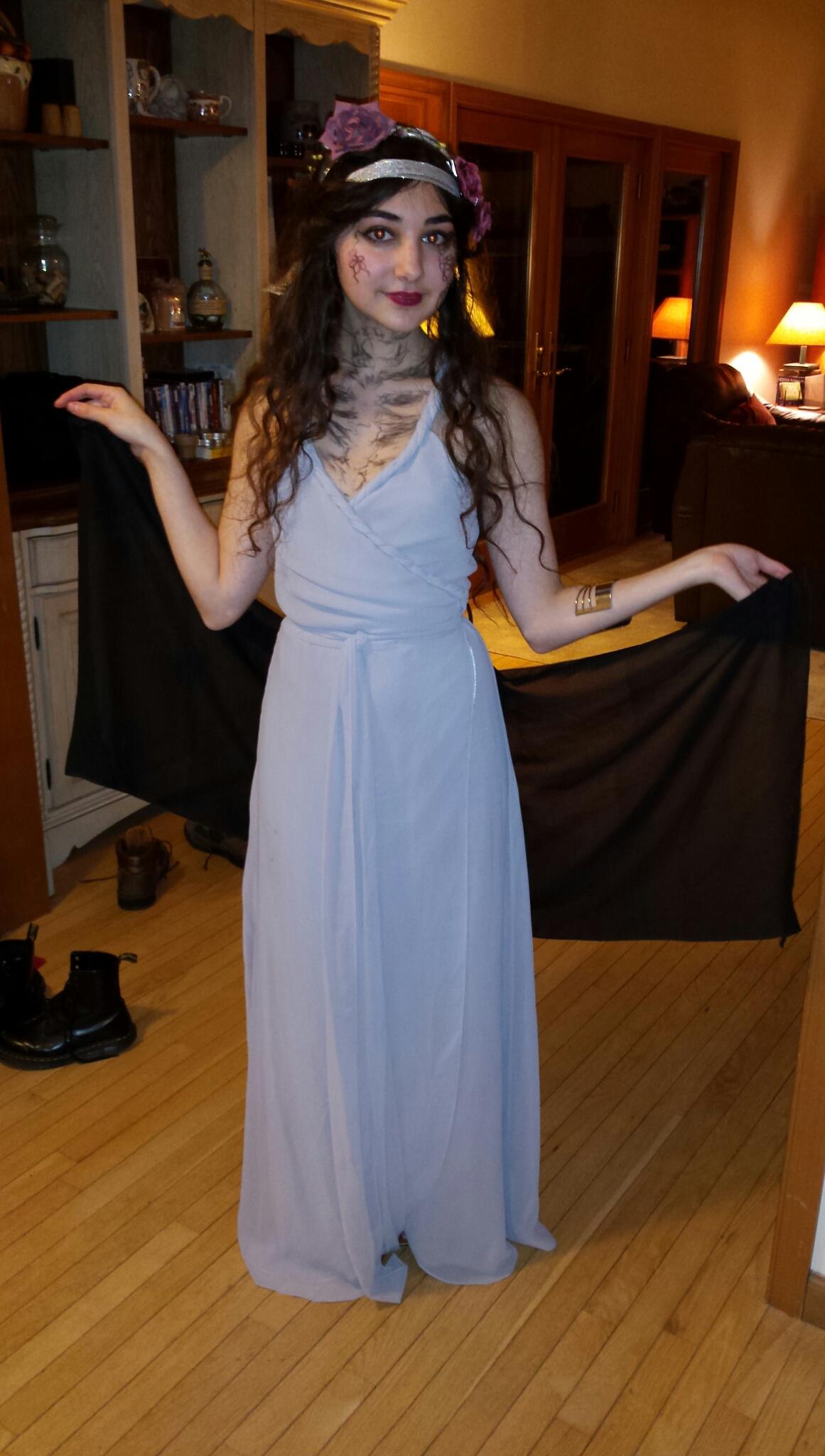 a young woman stands on a hardwood floor in a long, flowing grey wrap dress and a black shawl. she is wearing a floral crown and has dark makeup which gives a slightly spooky or gothic look. the setting appears to be a home, with wooden furniture and cabinetry visible in the background.  she looks mysterious and ethereal in her costume.