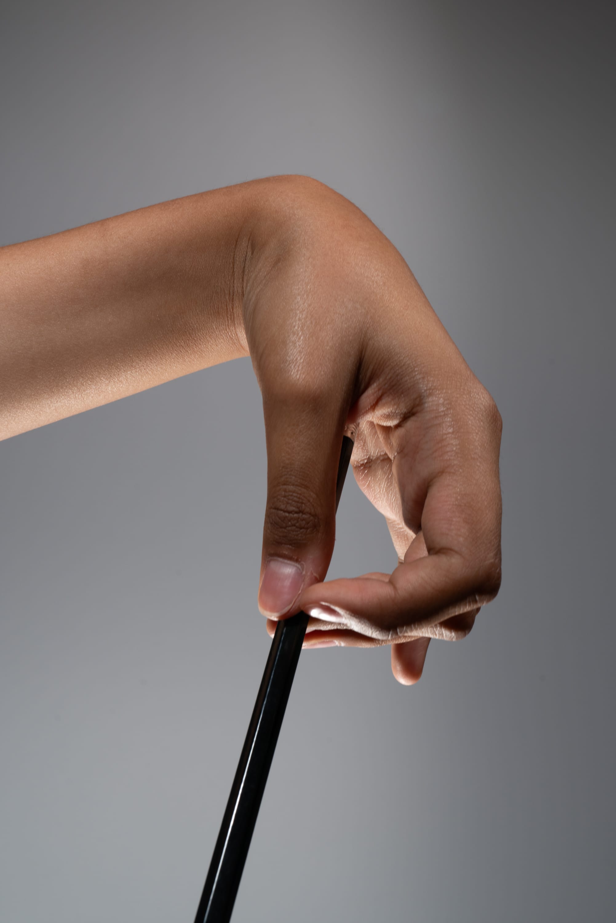 the image shows a closeup of a hand gripping a black pencil against a gray background. the hand is bent at the wrist, with the fingers curled around the pencil. the lighting is bright and even, highlighting the details of the hand and pencil. the overall composition emphasizes the strength and dexterity of the hand.