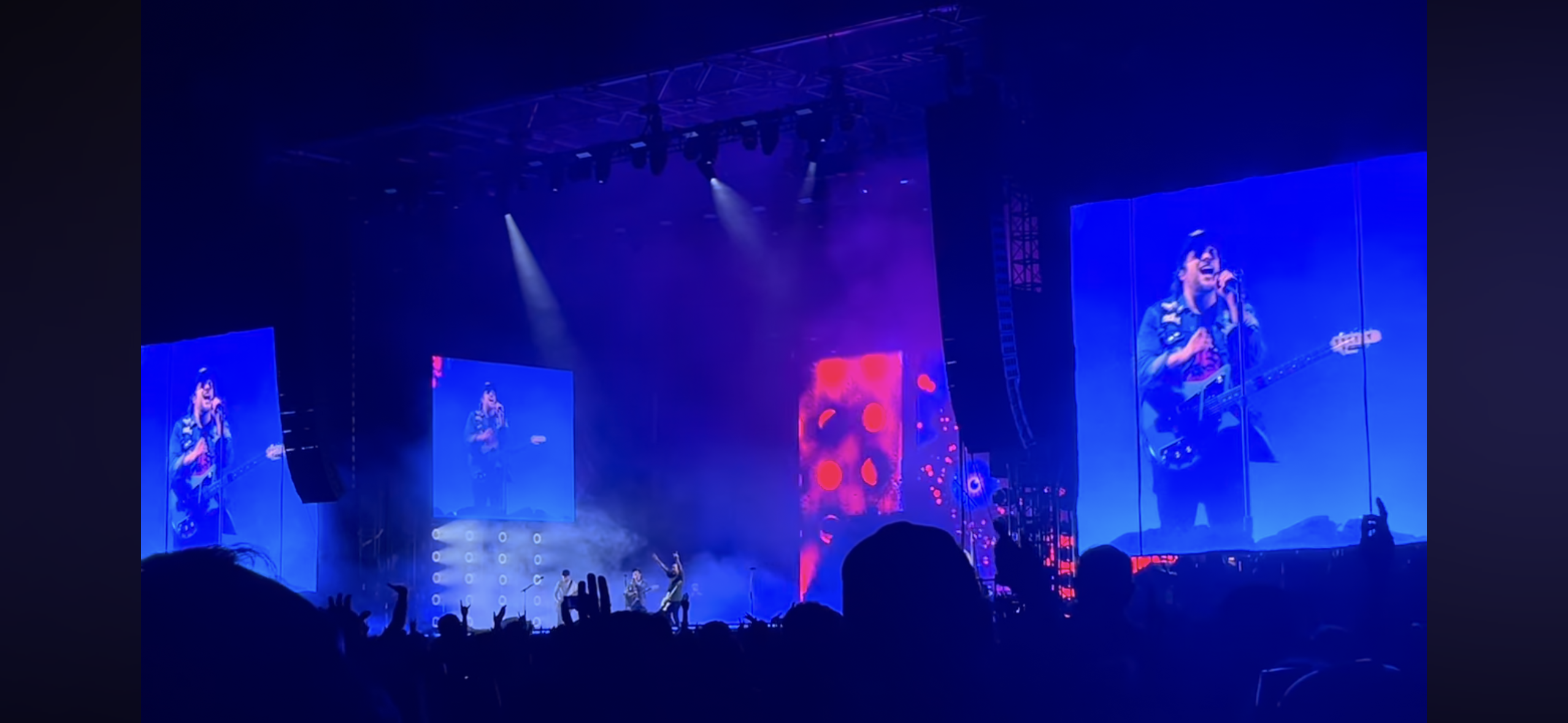 a concert scene with a large crowd of people watching a band perform on stage. the stage is lit up with bright spotlights and there are two large screens showing closeups of the band members. the band is playing energetic music and the crowd is singing along. the atmosphere is electric with excitement and energy.