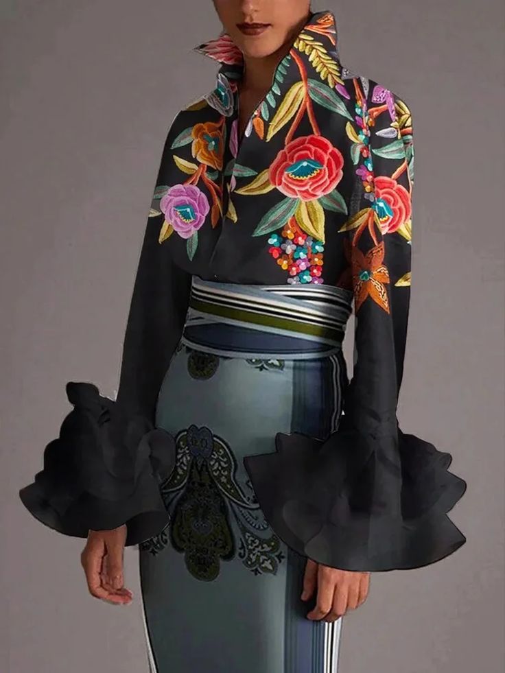 a woman is wearing a black dress with floral embroidery. the dress has large ruffled sleeves and a high collar. the woman is standing in front of a grey background.  she is wearing a black dress with a floral pattern and is posing for the camera. she has a beautiful smile and long hair. she is wearing a stunning outfit that is perfect for a special occasion.
