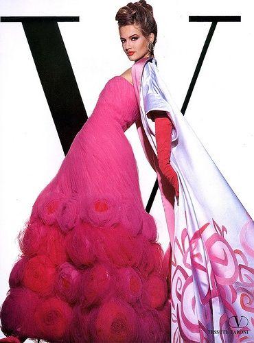 a studio shot features a model in a strapless pink dress and long red gloves with a white satin shawl with pink floral patterns on it. the dress transitions from pink to dark red. the model wears a bouffant hairstyle and large earrings. the image includes a large "v" in the background, which is part of the valentino brand identity. the dress skirt has roselike patterns all over it, and the bottom right corner includes the "tessuti yakont" label.