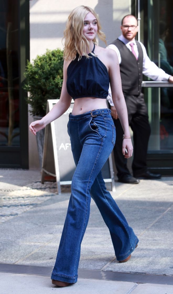 elle fanning is captured in a candid street style shot.  she's wearing a dark blue halter crop top paired with highwaisted, dark wash bellbottom jeans. her blonde hair is styled loosely. she's walking on a city sidewalk, passing by a building entrance where a man in a suit is visible in the background.  the overall vibe is casual summer fashion.