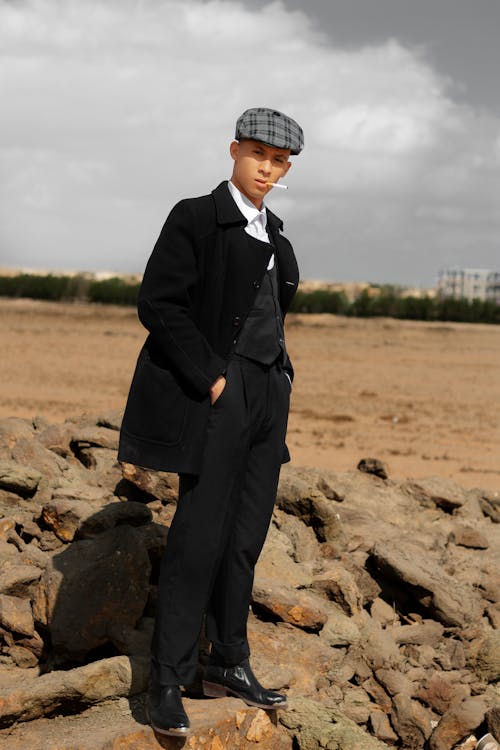 a young man in a black coat and pants stands on a rocky desert landscape. he is wearing a newsboy cap and boots. he has a cigarette in his mouth. the photo is styled in a retro or vintage fashion, with a slightly desaturated color scheme. the overall mood is serious and cool.