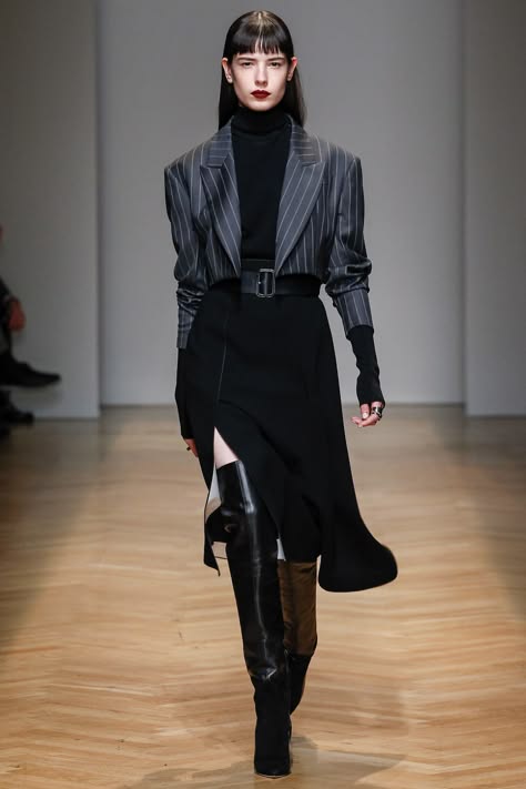 a female fashion model is walking down a runway wearing a stylish outfit. she is wearing a black pinstripe blazer over a black turtleneck sweater, paired with a black skirt and black kneehigh boots. she has a black belt with a silver buckle around her waist. she has black hair and red lips. she is striking a pose for the camera.  the runway is a wooden floor.