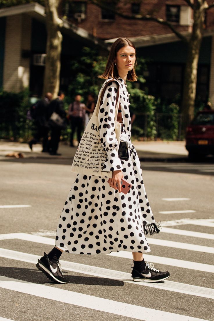 a stylish young woman in a black and white polka dot midi dress walks across a city street. she carries a tote bag with text on it and is wearing black nike sneakers. the image is a street style photograph taken during the daytime. the background is blurred, showcasing urban surroundings.  she appears confident and fashionable.