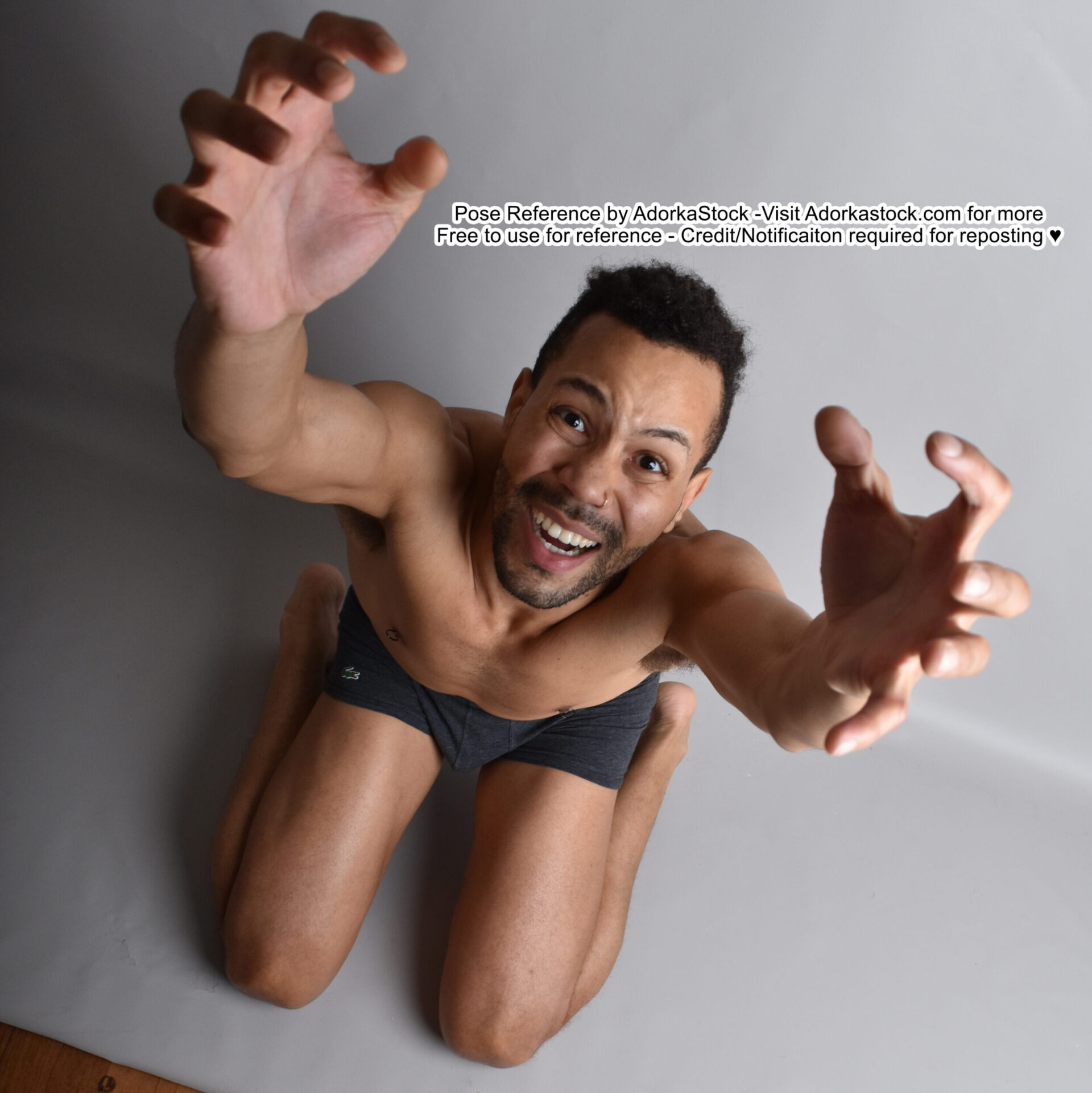 a man in his underwear is kneeling and looking directly up at the camera. he is laughing with his arms and hands outstretched toward the camera, his fingers are spread wide. he is muscular and has a toned physique. the man is in a studio setting, with a grey background.