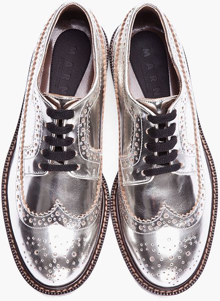 closeup, overhead shot showcases a pair of women's marni silver metallic leather brogue shoes. the shoes are wingtip oxfords with intricate perforated detailing along the toe cap and edges.  they have black laces and a slightly chunky sole. the marni logo is visible on the insole of each shoe. the image highlights the shoes' texture and reflective quality of the metallic leather.