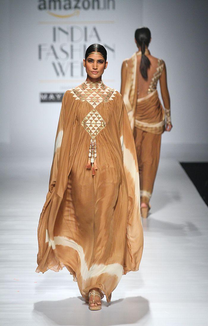 a woman in a brown dress walks the runway at a fashion show, another model is in the background wearing a sari. the background features logos of "amazon.in" and "india fashion week".