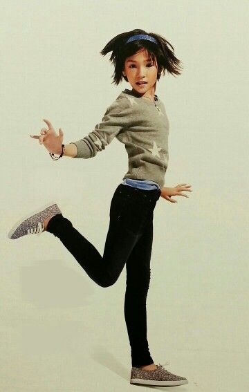 a young girl is captured in a fullbody shot against a plain, offwhite backdrop. she is joyfully leaping with her right leg raised and bent at the knee, giving the impression of midair suspension. she wears a light gray sweater adorned with white stars, paired with black leggings. her footwear consists of a pair of speckled sneakers. a blue headband keeps her short, dark hair off her face. she is turned to the right, extending both arms outwards, as if enjoying her moment.