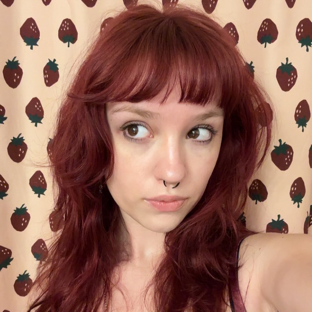 a selfie of a woman with red hair, bangs, and a nose ring, against a background of strawberries.