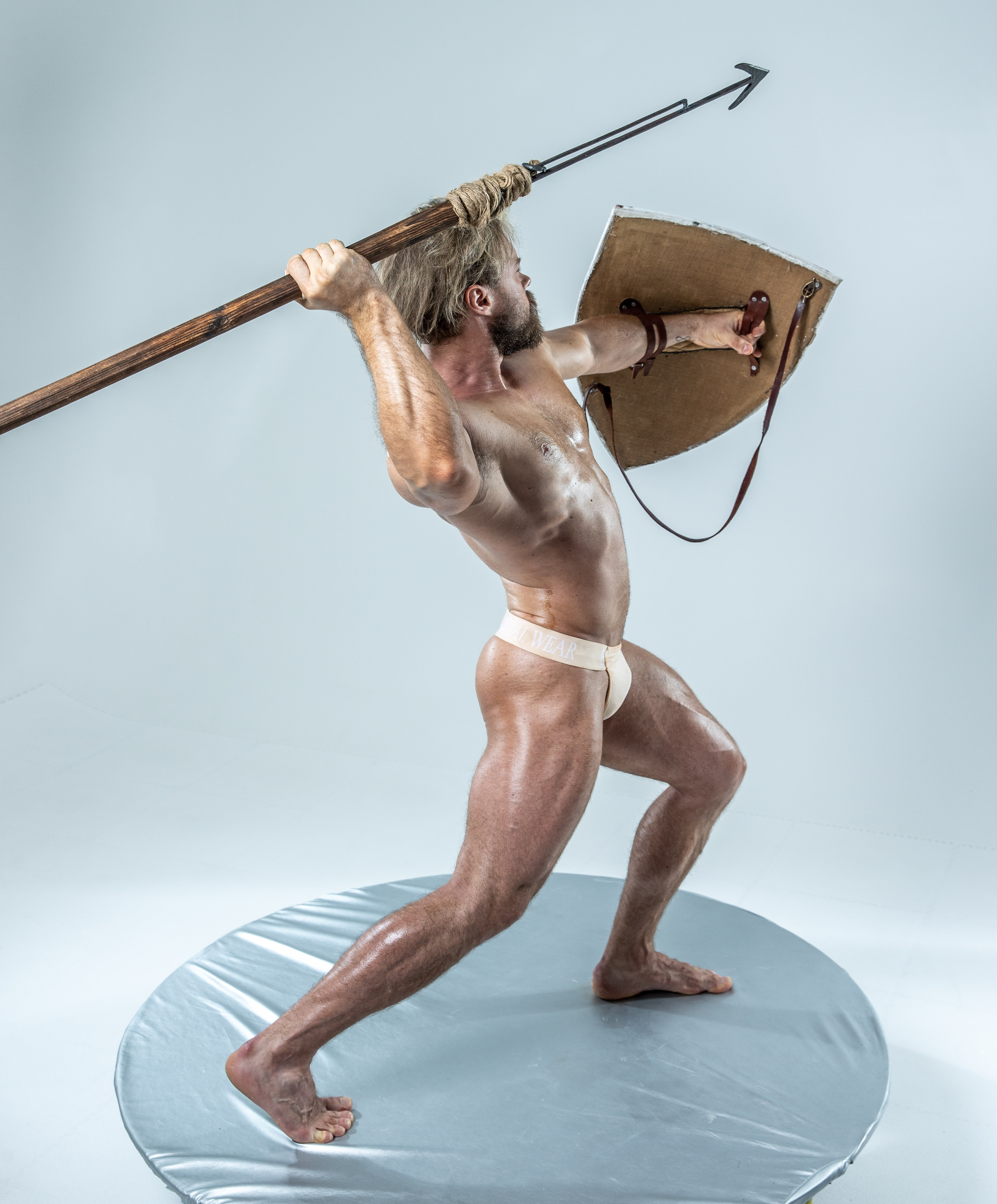 a muscular man with long blond hair in a warrior pose stands on a grey platform against a white background.  he holds a spear in his right hand and a shield in his left, as if ready for battle. his attire suggests an ancient greek or roman warrior. his body is glistening as if oiled.