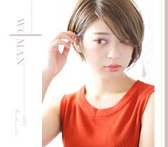 an asian woman with short brown hair styled in a bob cut, wearing a vibrant orange top. she is looking slightly down and to her left while lightly touching her hair. the background is a soft white with faint text overlay.