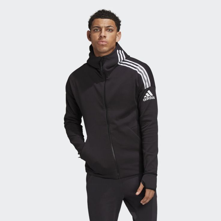 a man is modeling a black adidas hoodie with white stripes on the sleeve and a white adidas logo on the right side. he is standing with his arms relaxed at his sides. the hoodie has a zipper and a hood. the man is wearing black pants.