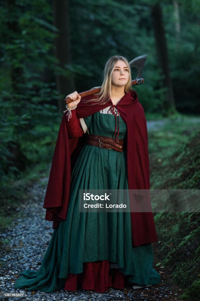 a full shot of a young blonde woman dressed in a medieval costume. she wears a long green dress with a red cloak and carries an axe on her shoulder.  she is standing on a path in a forest. the image has a fantasy and historical feel.