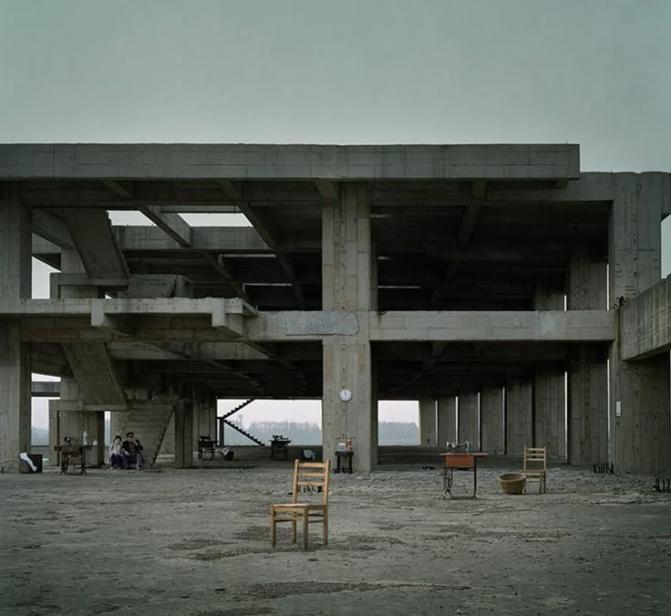 the photograph depicts a large, unfinished concrete building in a seemingly rural landscape. the structure is skeletal, with exposed beams and columns creating a gridlike pattern. it appears abandoned, with a sense of desolation.  the ground is bare earth and concrete.  a few wooden chairs and tables are sparsely scattered in the open space, along with a sewing machine. a small group of people are visible in the distance, adding a human scale to the vastness of the architecture. the overall tone is muted and grey, reinforcing the feeling of emptiness and abandonment. the sky is overcast, adding to the somber atmosphere. the style suggests minimalist architectural photography with a focus on the stark contrast between the manmade structure and the natural setting.