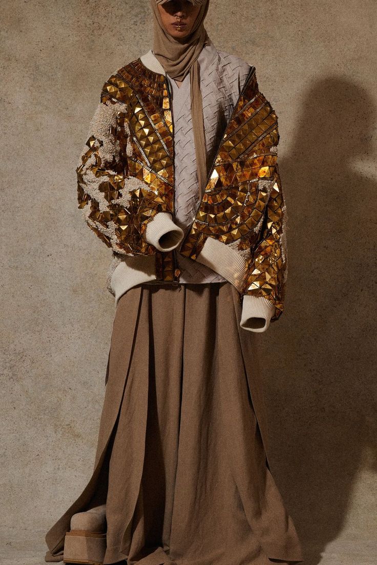 a woman is posing for the camera wearing a golden sequin jacket, a brown knitted scarf and a long brown skirt. she is wearing a hat and has a neutral expression on her face. the photo is taken in a studio with a neutral brown background.