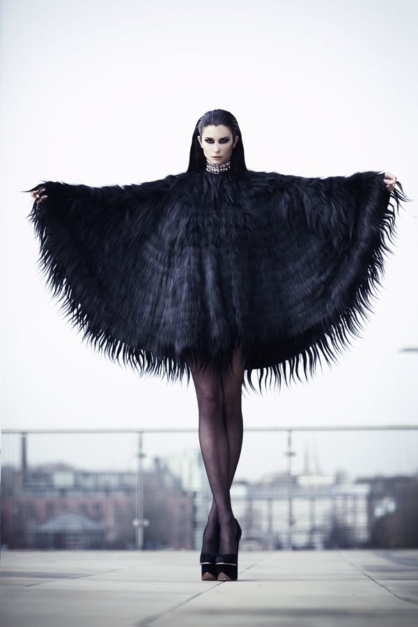a woman in a black fur cape and high heels stands with her arms outstretched,  posing for the camera in an urban setting.