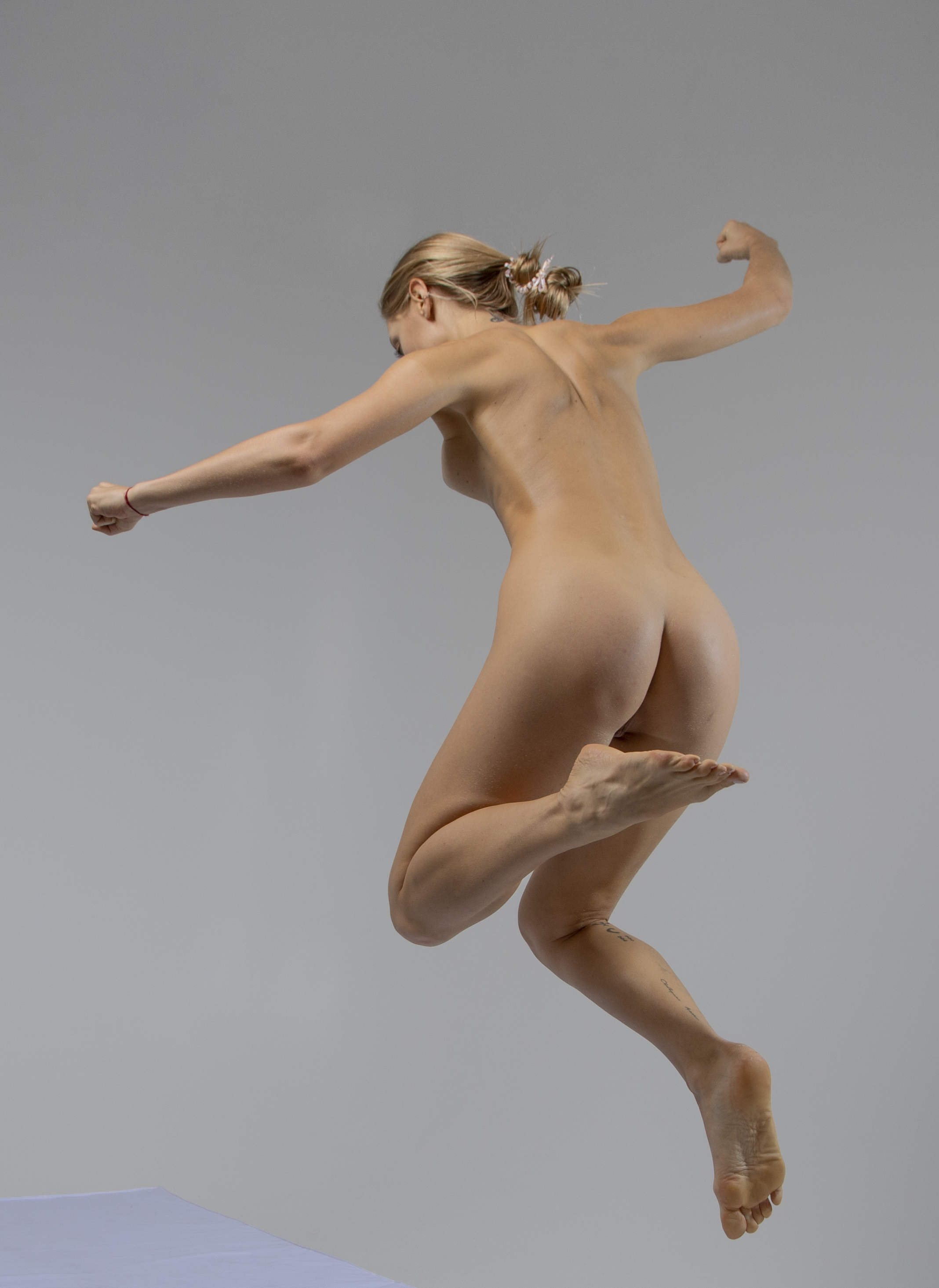 a full shot of a nude blonde woman in a dynamic pose, jumping barefoot in the air against a grey background. her arms are extended, fists clenched, and her ponytail is visible. the image captures her body in motion from a back view.