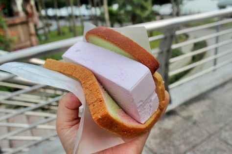 a closeup shot of a hand holding a delicious ice cream sandwich. the sandwich is made with two slices of bread, and in between, there is a large scoop of ice cream. the ice cream is a light pink color, and it looks very creamy and flavorful. the bread is lightly toasted, and it looks like it would be very crispy and satisfying to bite into. the whole sandwich looks very appetizing, and it would be a perfect treat on a hot day.
