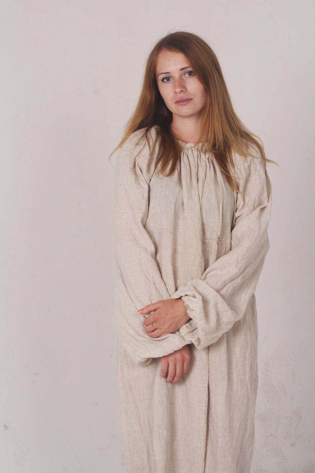 a woman in a long sleeve linen dress poses for a portrait in a studio. she is standing with her hands clasped in front of her.