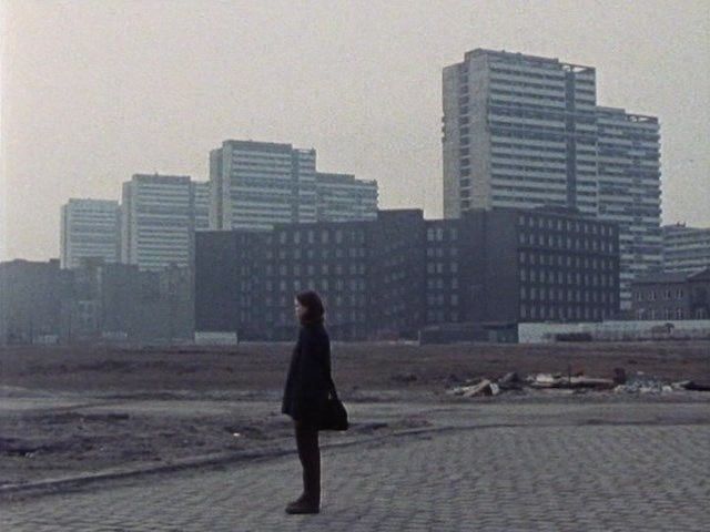 a still image from a film, possibly from the 1970s, depicts a woman standing alone in a desolate urban landscape. she is dressed in a dark coat and is facing away from the camera, looking towards a background of grey highrise apartment buildings. the foreground is a mostly bare, empty space that looks like a construction or demolition site. the overall mood is grey, lonely, and somewhat desolate, reflecting the bleakness of the urban environment.  the sky is overcast, and the overall color palette is muted and subdued, enhancing the film's atmosphere.