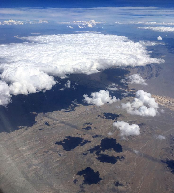 this aerial shot captures a dramatic landscape where fluffy white clouds cast dark shadows on a desert terrain. below the clouds, the terrain is a mix of browns and tans, marked with geological features possibly including mountains. the sky is a mix of blue and white, reflecting the overcast conditions. the photo's perspective gives a sweeping overview of the interplay between weather and the earth's surface.