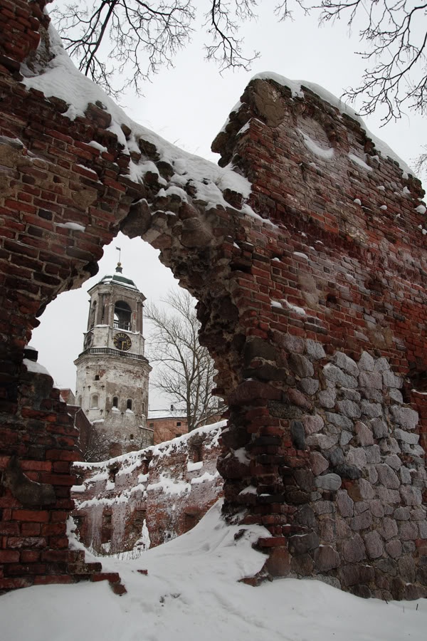 a photo of an old stone and brick castle in ruins. the castle is seen through an archway in the ruins, with a clock tower visible. there is snow on the ground and on the castle walls. the trees are bare, indicating it is winter.