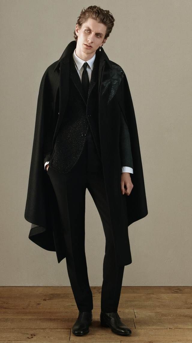 a fulllength portrait shows a man in a black suit and cape. he wears a white dress shirt and a black tie underneath the black suit jacket. the jacket has a decorated pattern on the left side. the cape is long and dramatic, with a high collar. the man stands on a light wooden floor, in front of a neutral wall. he wears black dress shoes.