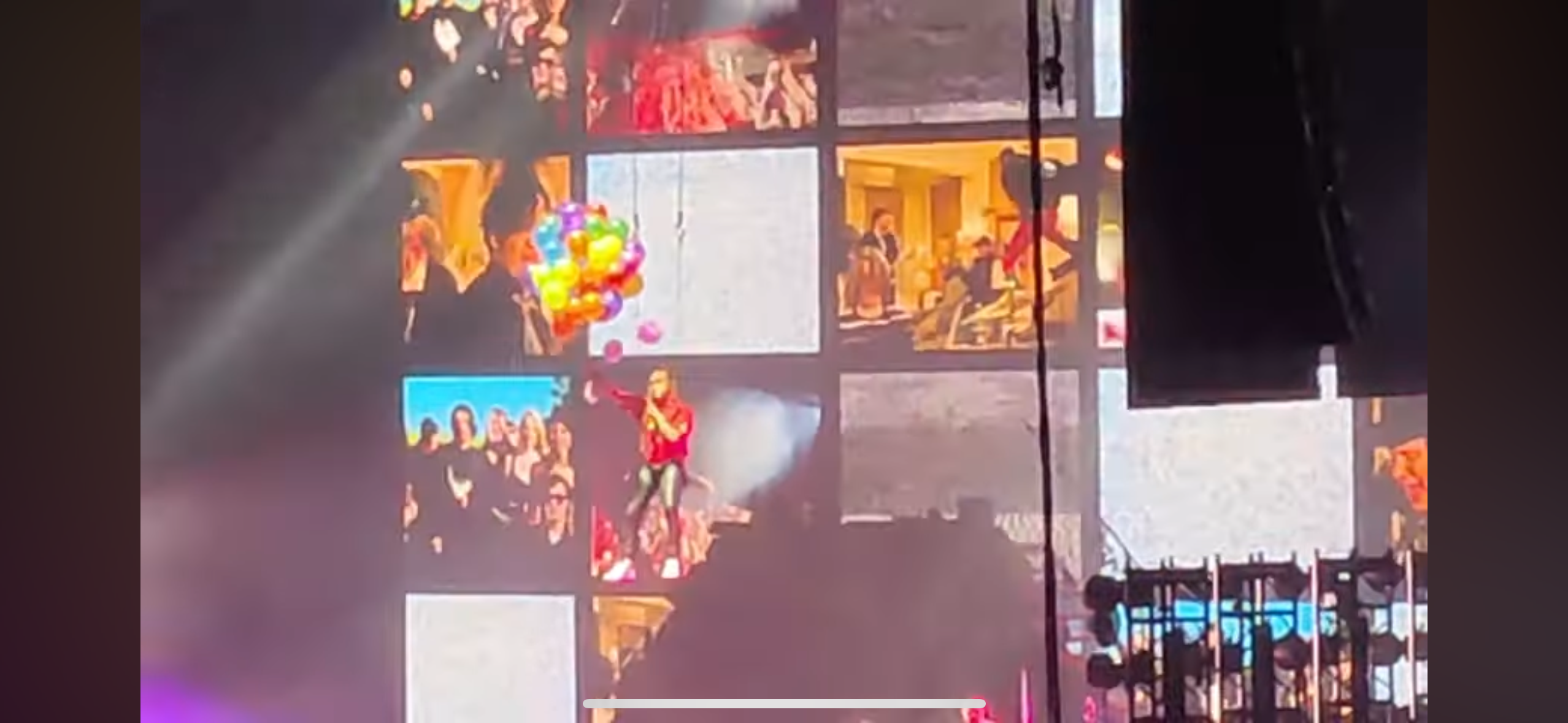 a concert stage with a performer standing in the middle of the stage. the performer is holding a microphone and is singing. there is an audience in the background and the stage is lit up with bright lights. there are also balloons and confetti in the air.