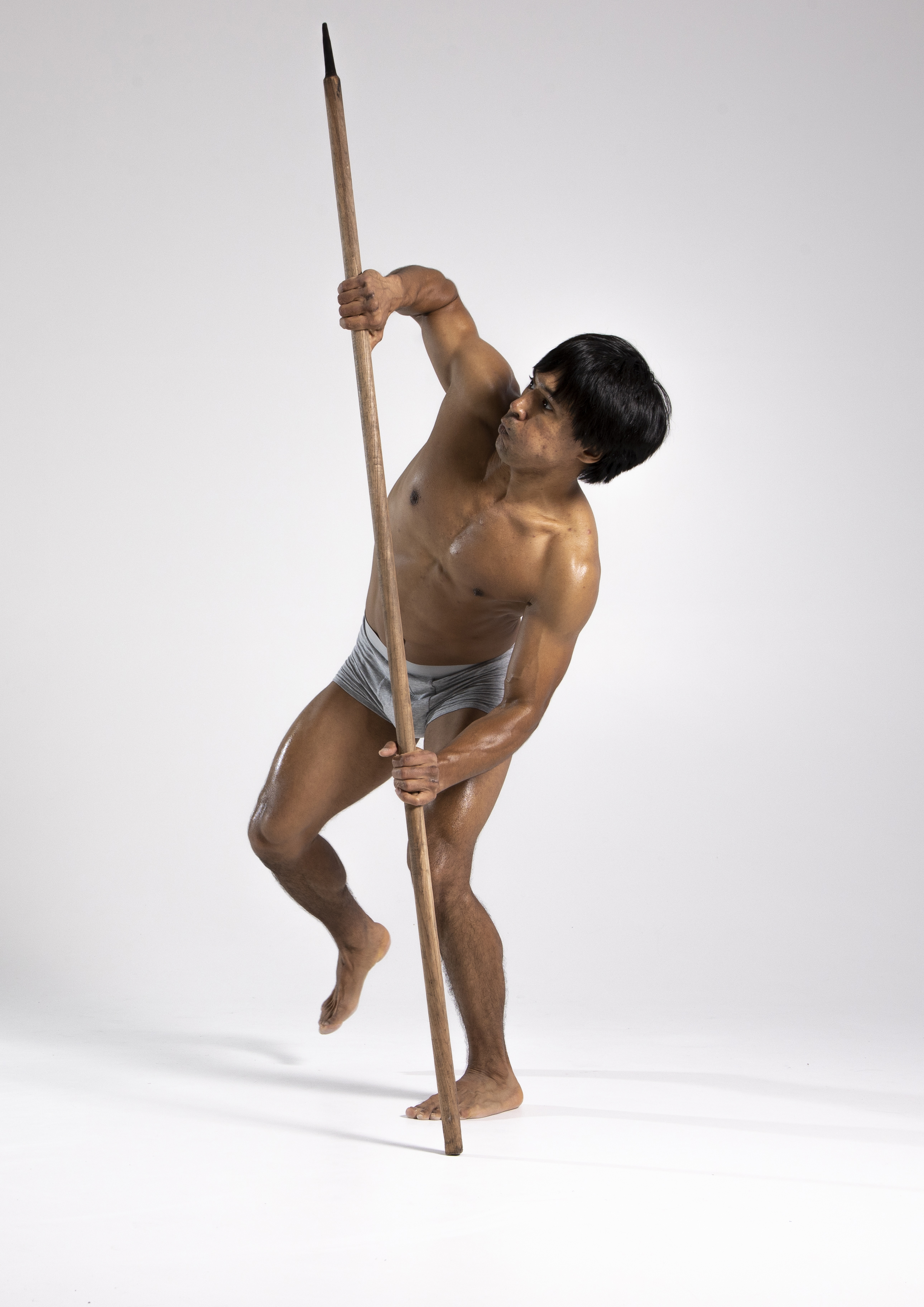 a barechested man in gray shorts stands in a studio setting, holding a spear. he is looking away with a muscular build and an athletic posture. his skin appears oiled or wet. the background is a plain, light color.