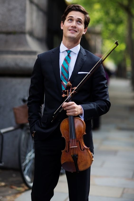 a young man in a sharp navy suit and striped tie stands on a city sidewalk, gazing upwards with a hopeful expression. he holds a violin in his left hand, its warm brown tones contrasting against his dark attire. a bicycle leans against a building in the background, suggesting a bustling urban setting bathed in the soft glow of natural light.