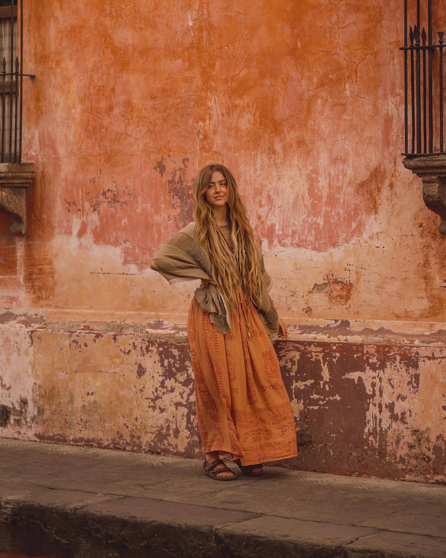 a fulllength outdoor portrait of a young woman with long blonde dreadlocks. she is wearing a flowing, orange maxi skirt with a subtle print, a tan shawl draped over her shoulders, and simple sandals. she stands against a backdrop of a weathered, orangetoned stucco wall with visible texture and peeling paint. the overall aesthetic is bohemian and suggests travel or exploration, possibly in a latin american setting. the image has a warm, natural light.