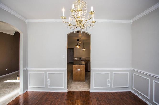 interior view of a dining room with hardwood floors, light gray walls with wainscoting, and a crystal chandelier hanging from the ceiling. an archway leads to a kitchen area with a central island and pendant lighting.