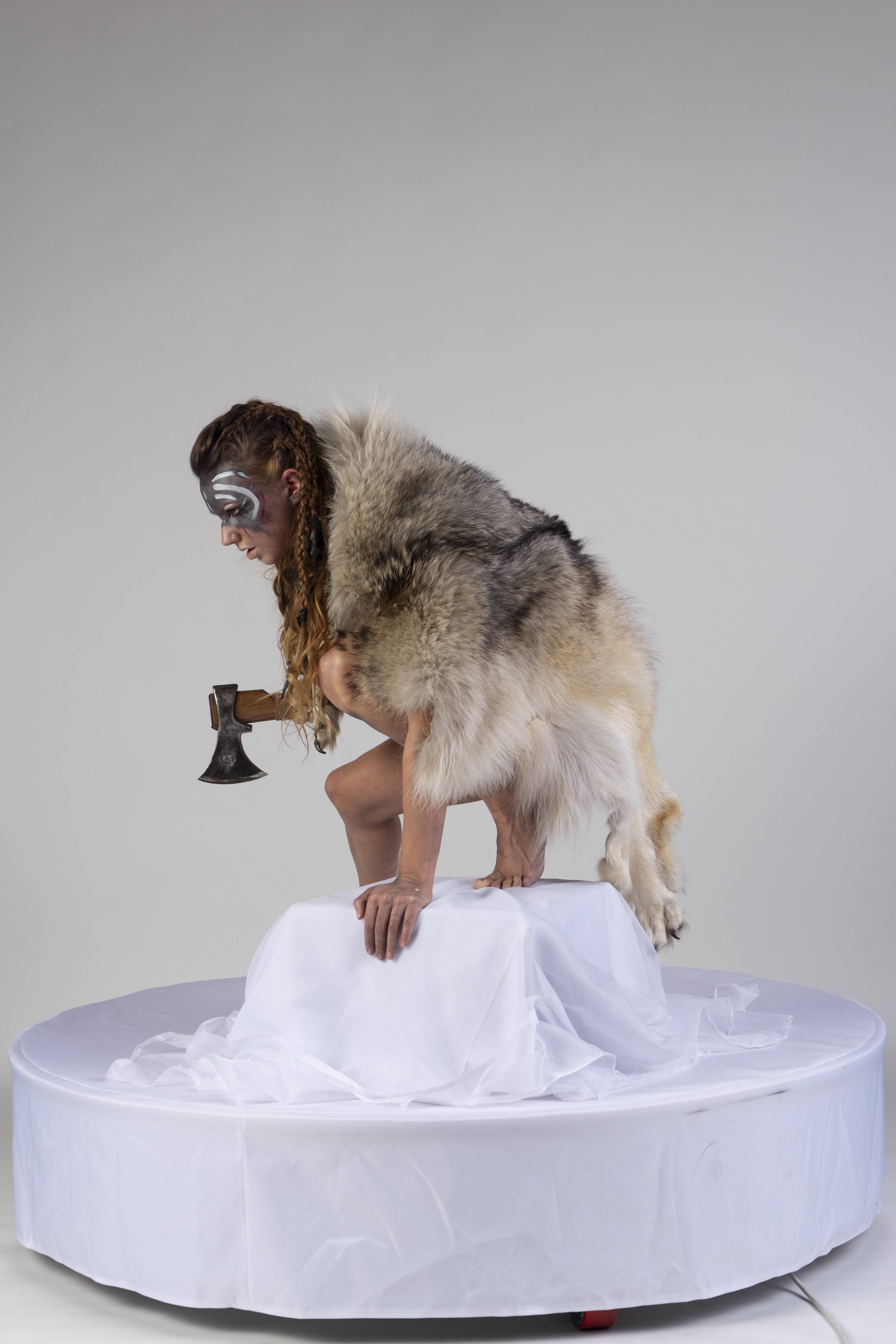 a woman in tribal garb with face paint stands on a white pedestal, holding an axe and draped in fur, set against a plain white background.