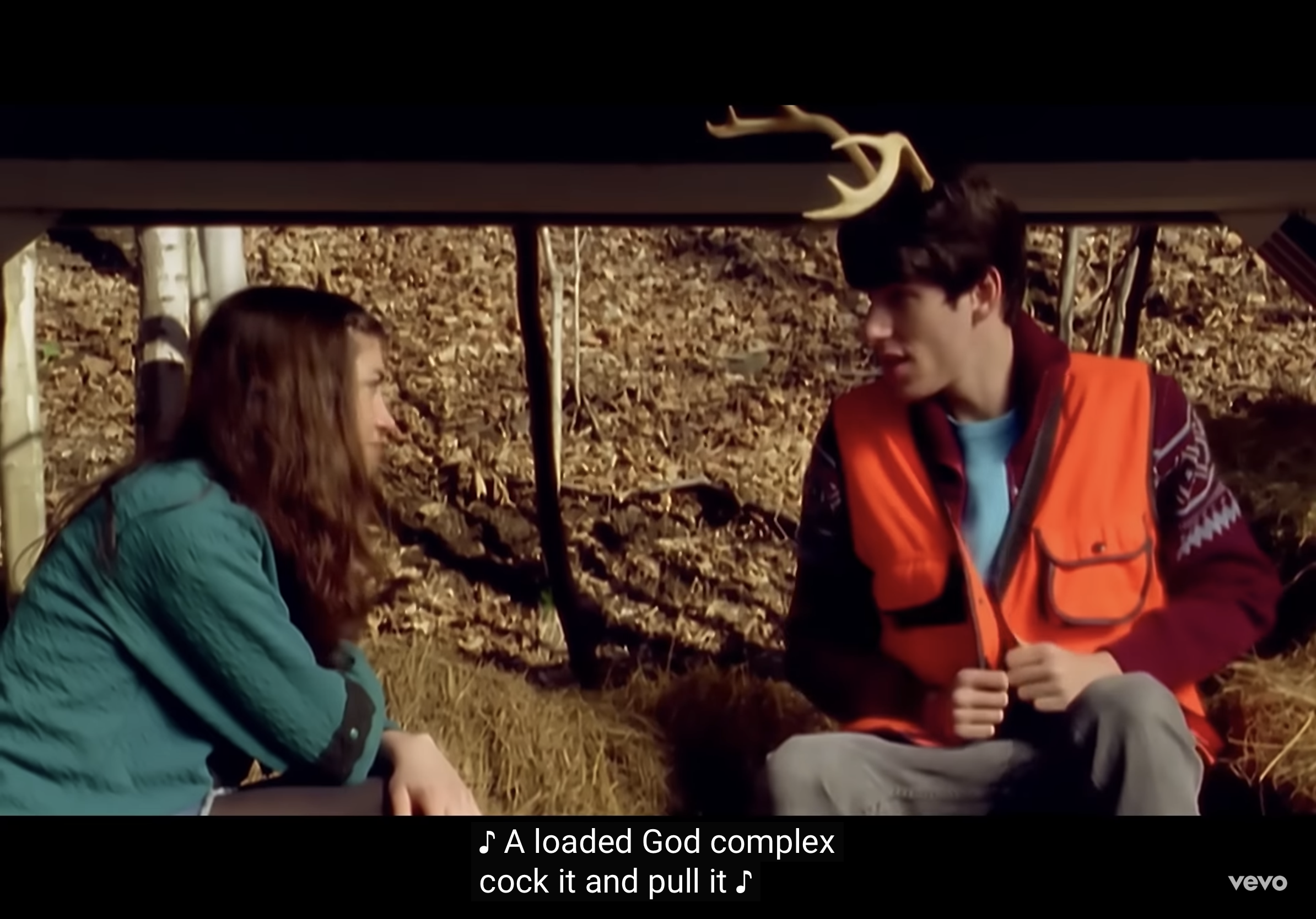 a young man and woman sit beneath a simple wooden structure.  he wears a bright orange hunting vest and deer antlers on his head.  they appear to be having a conversation, and the text overlaid on the image is a lyric from a song: "a loaded god complex, cock it and pull it." the background suggests they are in the woods.  the overall tone is both humorous and slightly unnerving.