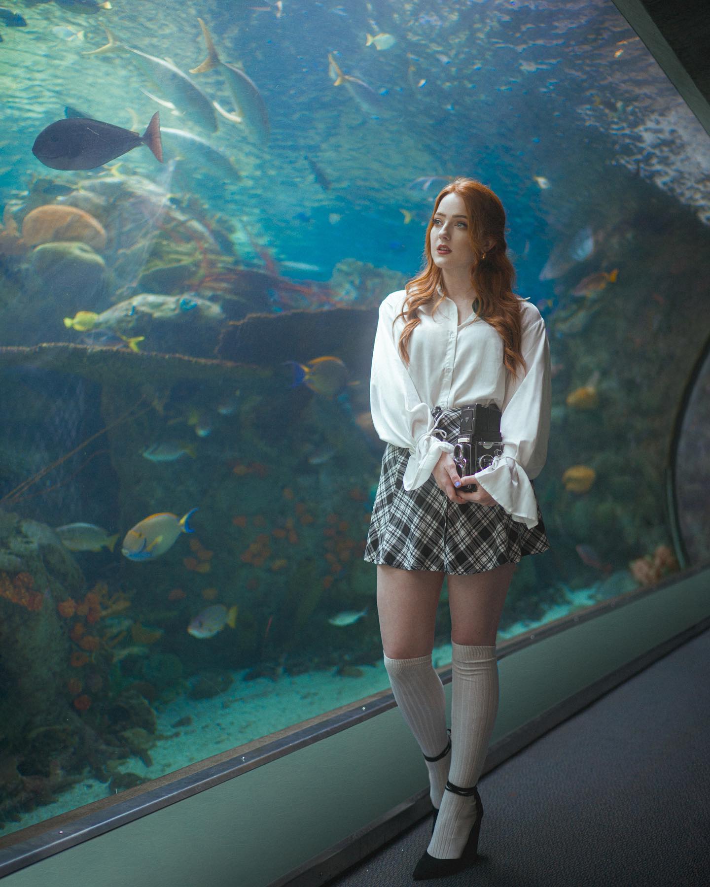 young woman with long red hair stands in an aquarium tunnel, gazing at the fish swimming past. she's wearing a white blouse, a black and white plaid skirt, white kneehigh socks, and black heels. she's holding a black clutch purse in her hands. the aquarium is brightly lit with natural light filtering in from above.