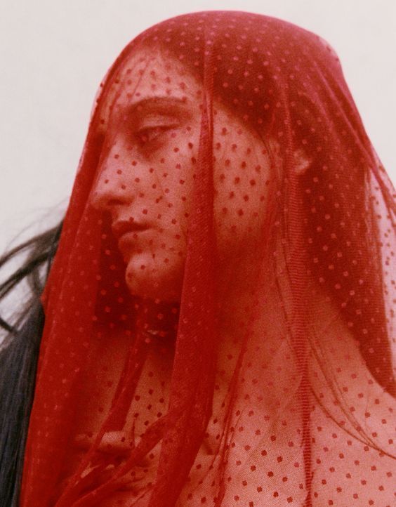 a woman is wearing a red veil with polka dots. the veil is covering most of her face, but her eyes are visible. the woman's expression is mysterious and sensual. the image is close up and the woman's hair is partially visible. the image is set against a white background and the veil is the