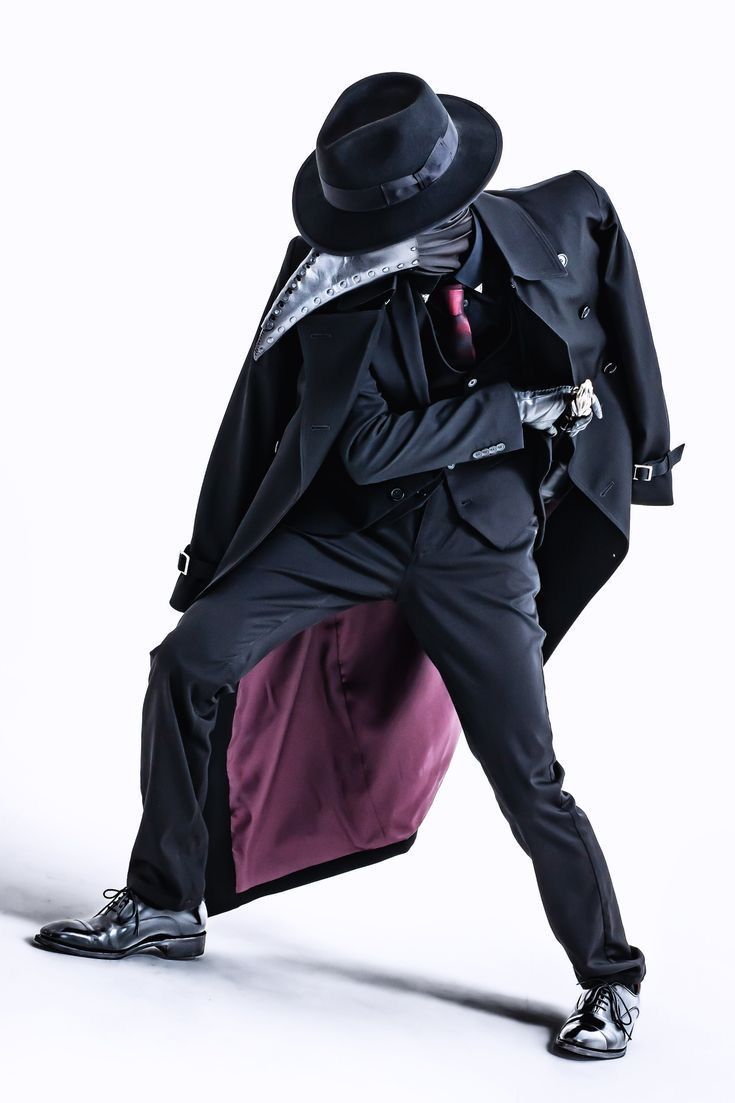 a man wearing a black suit with a long coat, black hat, gloves and a mask stands against a white background.