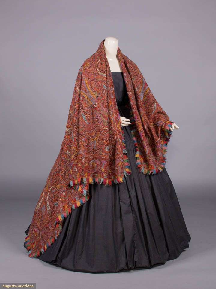 the image features a mannequin wearing a darkcolored dress with a large, paisleypatterned shawl draped over its shoulders and down its front. the shawl has a multicolored fringe along the bottom edge. the overall impression is of historical or vintage fashion, possibly from the victorian era, displayed in a professional setting.