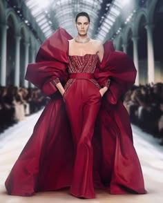 a fashion model walks the runway in a stunning red gown with dramatic puffed sleeves. the gown is a classic haute couture design, with a flowing skirt and a fitted bodice adorned with sparkling embellishments. the model is wearing a simple necklace and her hair is styled in a sleek updo. she is posing confidently for the photographers, and her look is both elegant and glamorous.