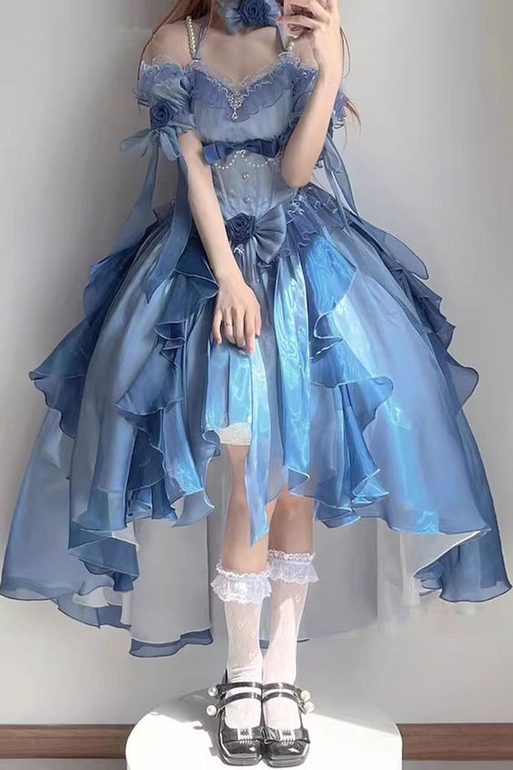 a young woman with long brown hair in a beautiful blue lolita dress with ruffles. she is posing with a white background. the dress is very detailed with lace and bows, and it also has a train. she is wearing white lace socks and white shoes, which are also very detailed.  the overall look is very elegant and sweet.