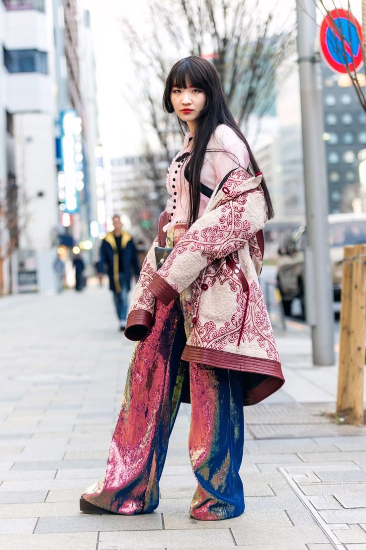 a stylish asian woman stands on a city sidewalk wearing eyecatching fashion. she sports wideleg sequin pants with a redtoblue gradient, a pink buttonup shirt, and an intricately patterned coat draped over her shoulders. her dark hair features blunt bangs, and she wears bold red lipstick. the urban background includes buildings and a street sign.