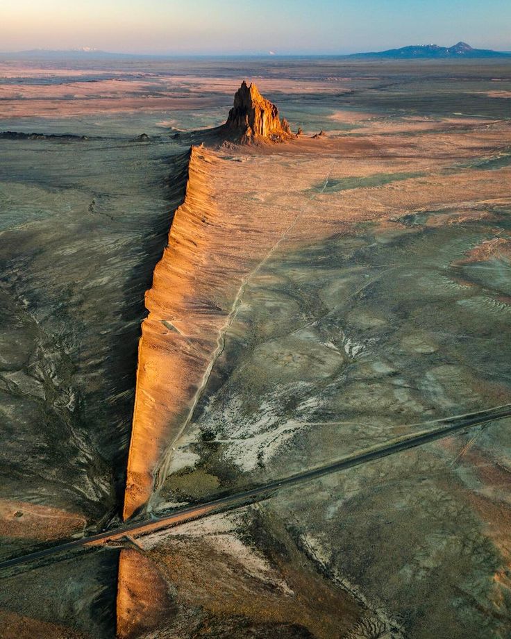 an aerial view of a rock formation in the desert. the rock formation is a tall, pointed peak that rises up from the desert floor. it is surrounded by a vast expanse of dry, brown land. the sun is setting in the distance, casting a long shadow over the rock formation. the image was taken from a high vantage point, which gives a sense of the scale of the rock formation and the surrounding desert.