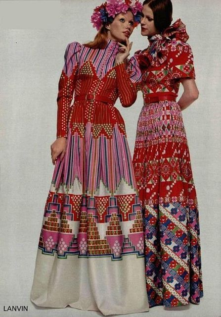 this image shows a vintage advertisement for lanvin featuring two models wearing vibrant, long dresses. the dresses are predominantly red, with pink and other colors in intricate geometric and floral patterns.  the dresses are maxi length, aline and feature long sleeves, belted waists, and a pleated skirt effect. one model wears a flower crown. the overall style is distinctly 1970s bohemian chic. the image is set against a simple light background.